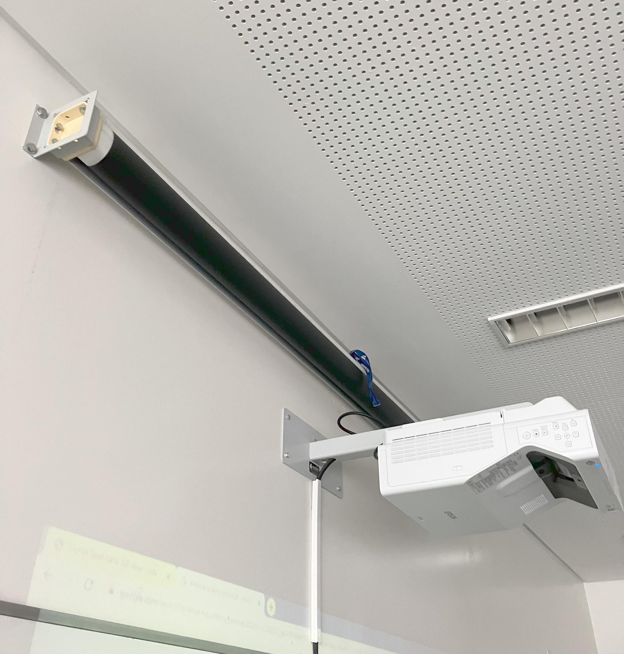 Een projector hangt aan een muur. Een scherm hangt er opgerold boven. Het scherm kan zo niet uitgerold worden, en de projector kan er niet op projecteren.